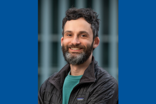 Pictured is Dr. Jesse Shapiro, a man with dark wavy hair and a beard, smiling at the camera. He is wearing a teal sweatshirt and a black Patagonia jacket. The background is blurred with vertical blue-green structures.